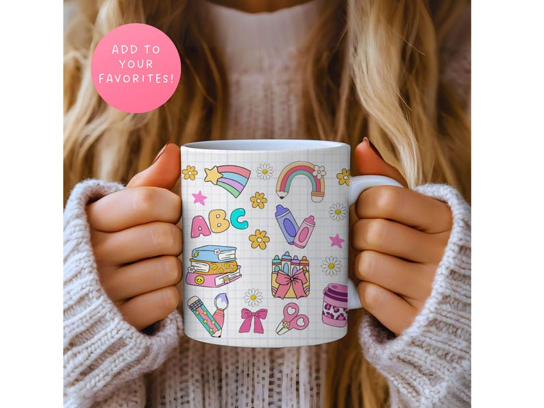 May include: White ceramic mug decorated with colorful school-themed illustrations, including a rainbow, crayons, books, and the letters "ABC". The mug is held by a person wearing a beige sweater. A pink circle with the text "ADD TO YOUR FAVORITES!" is in the background.