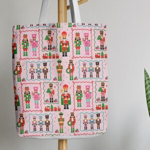 May include: A pink tote bag featuring a repeating pattern of colorful nutcracker soldiers, toy trains, and Christmas trees within decorative frames. The bag has white straps and is hanging on a wooden rack.