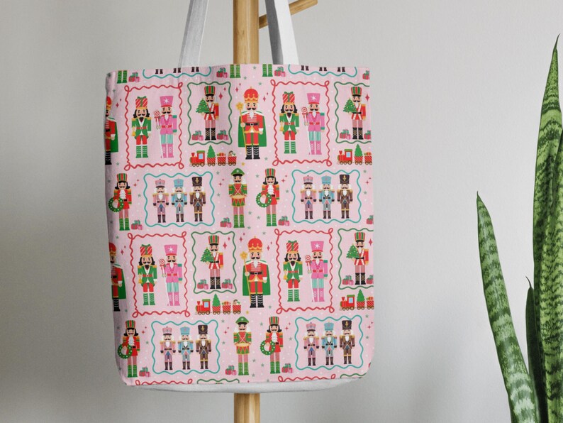 May include: A pink tote bag featuring a repeating pattern of colorful nutcracker soldiers, toy trains, and Christmas trees within decorative frames. The bag has white straps and is hanging on a wooden rack.