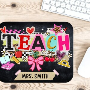 May include: Black mousepad with the word "TEACH" in colorful letters and teacher-themed graphics. Includes a pink bow, apple, pencil, and the name "MRS. SMITH". A white mouse and keyboard are in the background.