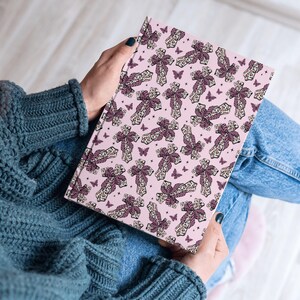 May include: A pink notebook with a repeating pattern of crosses and butterflies. The crosses are dark purple with a cream lace design. The butterflies are also dark purple. The notebook is being held.