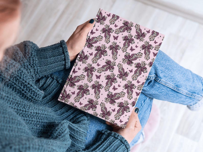 May include: A pink notebook with a repeating pattern of crosses and butterflies. The crosses are dark purple with a cream lace design. The butterflies are also dark purple. The notebook is being held.