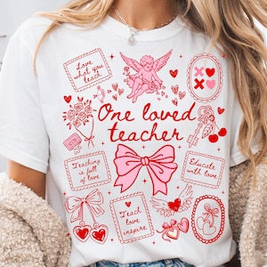 Puede incluir: Camiseta blanca con un diseño gráfico rojo y rosa. El diseño incluye el texto "One loved teacher" con ilustraciones de corazones, un lazo, un ángel y las frases "Love what you teach" y "Teach love inspire".