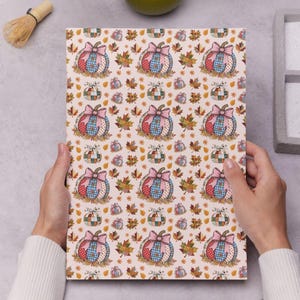 May include: A patterned paper featuring a repeating design of pumpkins with pink bows, surrounded by autumn leaves. The pumpkins have a patchwork design in red, blue, and tan. The background is a light beige color.