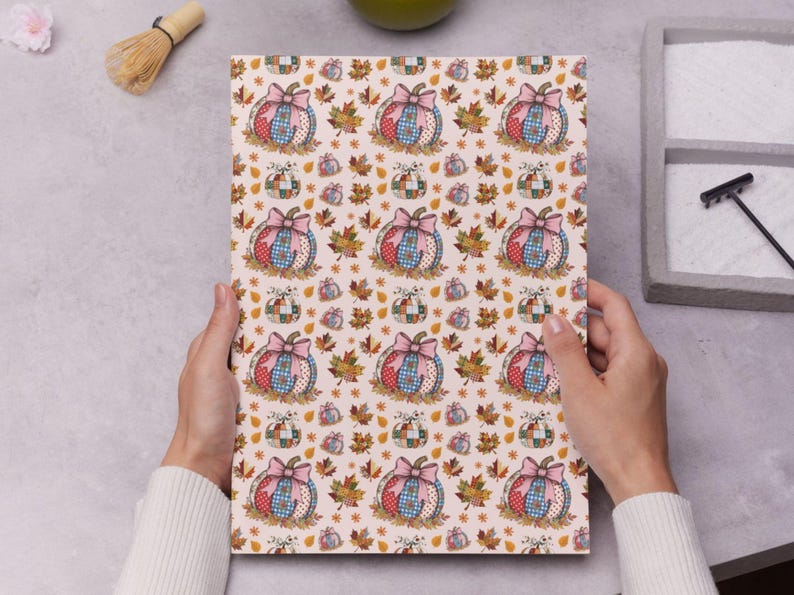 May include: A patterned paper featuring a repeating design of pumpkins with pink bows, surrounded by autumn leaves. The pumpkins have a patchwork design in red, blue, and tan. The background is a light beige color.