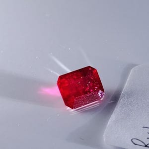 May include: A faceted, deep red gemstone with a crystalline structure. The gem is rectangular with beveled edges, reflecting light. The stone is set against a light gray background.