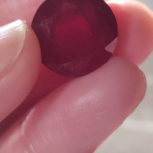 May include: A faceted, round, deep red gemstone is held between fingers. The gem has a polished surface and a slight sparkle, reflecting light. The color is a rich, dark shade of red.