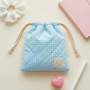 May include: A light blue and white gingham drawstring pouch with diagonal quilting. A small rectangular label on the front features a blue heart outline and the text "Bit & Thread". The pouch has beige rope drawstrings on both sides.