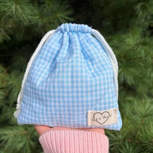 May include: A light blue and white gingham drawstring pouch with a quilted texture and white rope cords. A small rectangular fabric label is sewn onto the lower right corner, featuring a black heart outline and the text Rak & Thread.