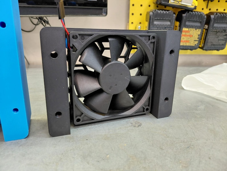 May include: A black computer fan with a protective frame. The fan has multiple blades and is mounted on a black bracket. Wires extend from the fan.