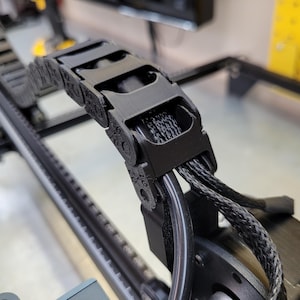 Op de afbeelding: Zwarte kabeldrager met een reeks scharnierende schakels, die meerdere zwarte kabels vasthouden. De drager is gemonteerd op een metalen rail, met een motor zichtbaar. Ontwerp voor industriële of robottoepassingen. Afmetingen in centimeters.