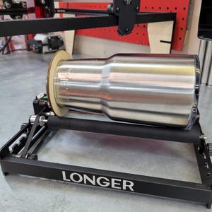 Puede incluir: Una máquina de rotación de vasos de metal negro con un vaso de acero inoxidable en una plataforma giratoria. La máquina tiene una base negra con la palabra "LONGER" impresa en ella.