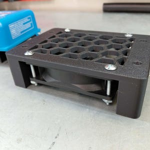 May include: A black electronic component with a honeycomb-patterned top and a fan. The component is secured with silver screws and has a rectangular shape. A blue electronic device is visible in the background.