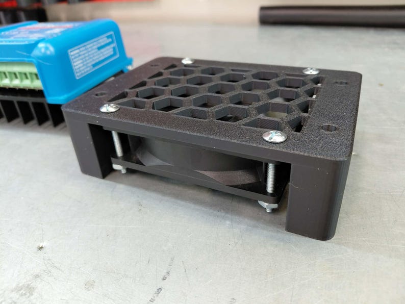 May include: A black electronic component with a honeycomb-patterned top and a fan. The component is secured with silver screws and has a rectangular shape. A blue electronic device is visible in the background.