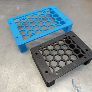 May include: Two 3D-printed rectangular frames with a honeycomb pattern. One frame is bright blue, and the other is black. Both have small feet and mounting holes. The frames are likely for electronics or a similar application.
