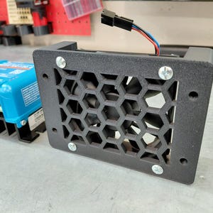 May include: A black metal fan with a honeycomb grill is secured with four silver screws. The fan is connected to a blue electronic device with a black heat sink. Wires with red, blue, and black insulation extend from the fan.