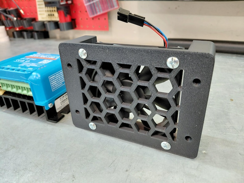 May include: A black metal fan with a honeycomb grill is secured with four silver screws. The fan is connected to a blue electronic device with a black heat sink. Wires with red, blue, and black insulation extend from the fan.