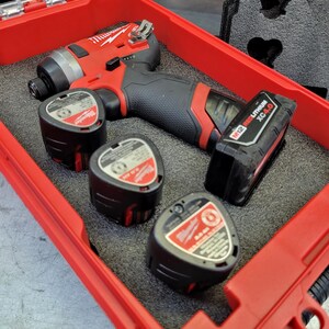 Op de afbeelding: Een rode en zwarte draadloze slagschroevendraaier met drie zwarte batterijpakketten en één bevestigd batterijpakket, allemaal in een rode koffer. De slagschroevendraaier heeft een zwarte grip en de merknaam "Milwaukee" is zichtbaar.