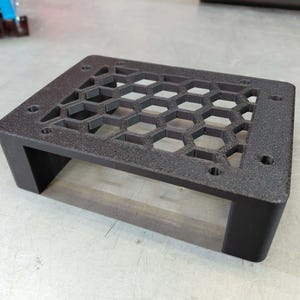 May include: A black, rectangular electronic component enclosure with a honeycomb pattern on top. The enclosure has four legs and several holes for mounting. The surface has a textured finish.