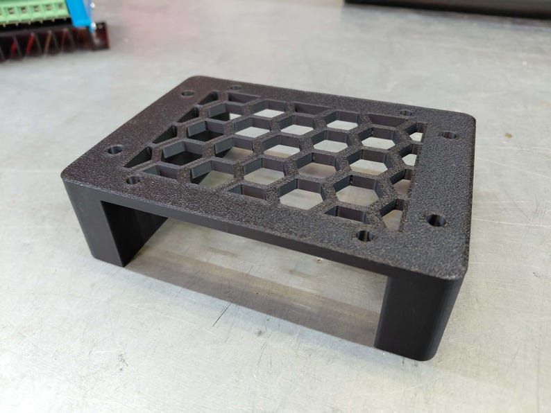 May include: A black, rectangular electronic component enclosure with a honeycomb pattern on top. The enclosure has four legs and several holes for mounting. The surface has a textured finish.