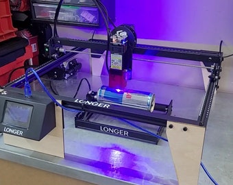May include: A black laser engraver with a red laser beam pointed at a silver can with the word "Red Bull" on it. The engraver has a blue cable connected to a black control panel with the word "LONGER" on it. The engraver is on a wooden table with a purple light shining on it.