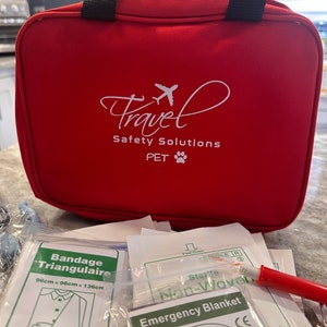 May include: Red travel first aid kit with black handles and the text "Travel Safety Solutions PET" in white. The kit is open and contains various first aid supplies, including a triangular bandage, sterile non-woven pads, and an emergency blanket.
