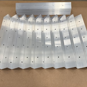 May include: A collection of silver-colored metal pieces, each with a series of evenly spaced holes. The pieces are arranged in a zig-zag pattern, with a separate stack of similar pieces in the background. The metal has a brushed finish.