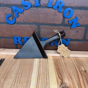 May include: A small, black cast iron spatula with a triangular blade and a black handle. The spatula is sitting on a wooden surface with a small brown tag that says "lol cray 12-21".