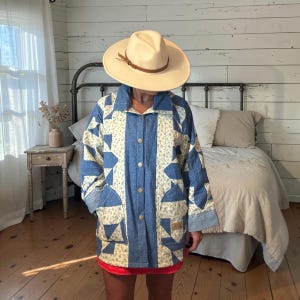 May include: A long-sleeved jacket featuring a patchwork design of blue denim and cream-colored fabric with a small floral pattern. The jacket has a button-up front, a collar, and two front pockets. A wide-brimmed tan hat with a brown band is worn.