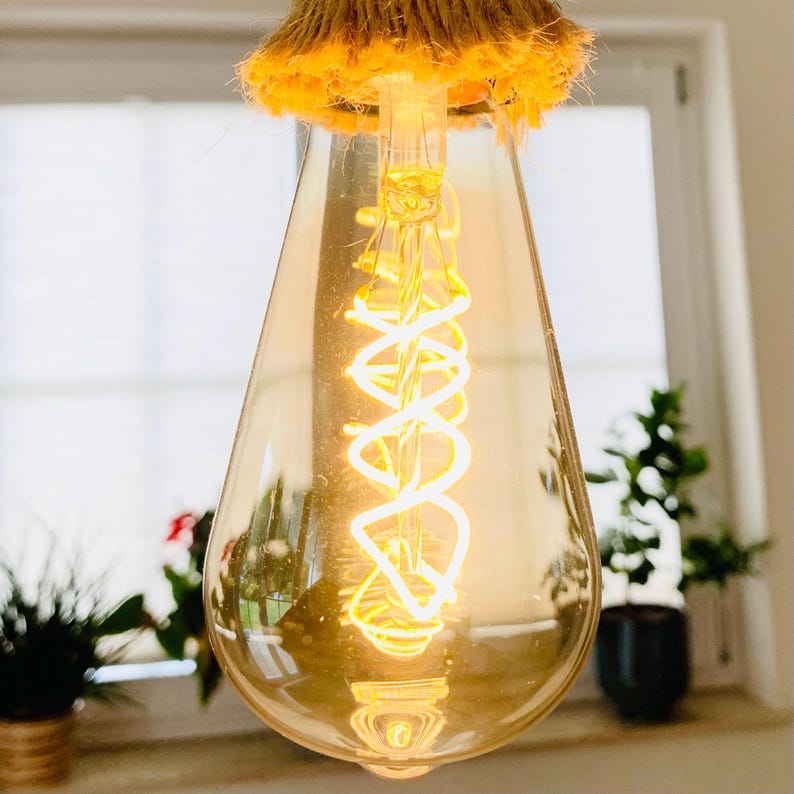 May include: A pear-shaped Edison light bulb with a warm, amber-tinted glass casing. Inside, a bright, glowing LED filament is arranged in a vertical spiral pattern. The top of the bulb features a textured, tan-coloured rope or twine wrapping.