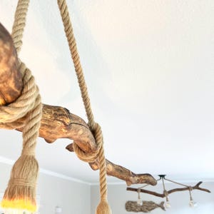 May include: A ceiling light fitting featuring a natural brown wooden branch suspended by thick tan jute ropes. The ropes are wrapped around the branch and terminate in light bulb sockets. A second similar branch fitting is visible in the background.