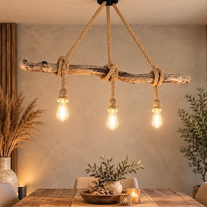 May include: A hanging light fixture featuring a horizontal piece of weathered driftwood suspended by thick, tan-coloured rope. Three Edison-style light bulbs hang from the driftwood, each secured by a matching rope wrap and socket.