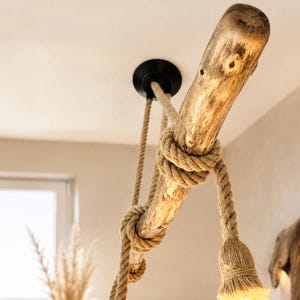 May include: A ceiling-mounted light fitting featuring a natural, light-brown wooden branch suspended by thick, tan-coloured twisted rope. The rope wraps around the branch and terminates in a light socket, creating a rustic, organic lighting design.