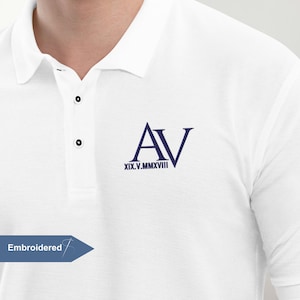 Könnte beinhalten: Weißes Poloshirt mit marineblauem, gesticktem "AV" Monogramm und römischen Ziffern. Das Shirt hat einen klassischen Kragen, drei Knöpfe und kurze Ärmel. Das Wort "Gestickt" steht in einer blauen Box.