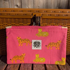 May include: Pink zippered pouch with a repeating pattern of yellow tigers and leopards. The pouch has a white label with the text "wildly loved" in black.