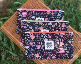 May include: Three black zippered pouches with a colorful heart and paw print pattern. Each pouch has a different colored zipper: pink, green, and orange. A white label with a heart design and the text "wholly loved" is on each pouch.