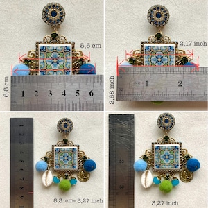 May include: A pair of blue and green ceramic tile earrings with gold accents and blue pom-poms. The earrings measure 2.68 inches in length and 2.17 inches in width. The tile is 1.77 inches wide and 1.77 inches tall.