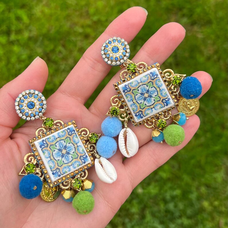 May include: A pair of blue and green statement earrings with a tile design, blue and green pom poms, and white seashells.