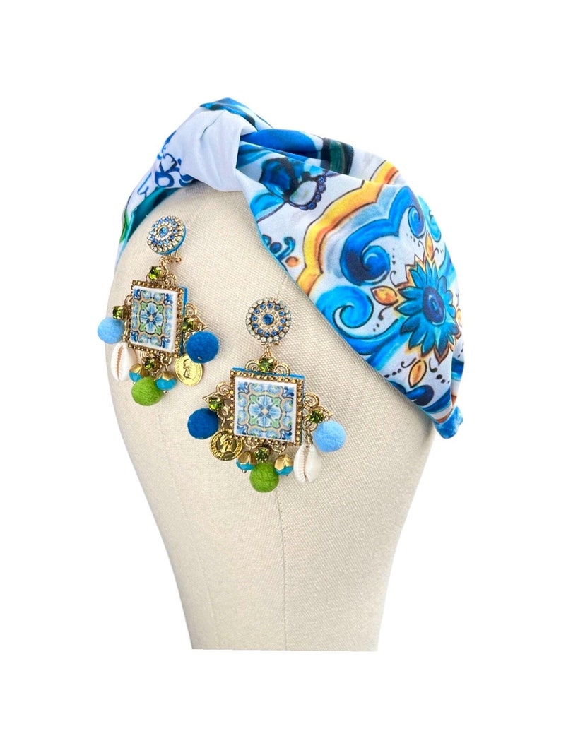 May include: A pair of blue and white ceramic tile earrings with gold accents and green and blue pom-poms. The earrings are hanging from a white and blue patterned headband.