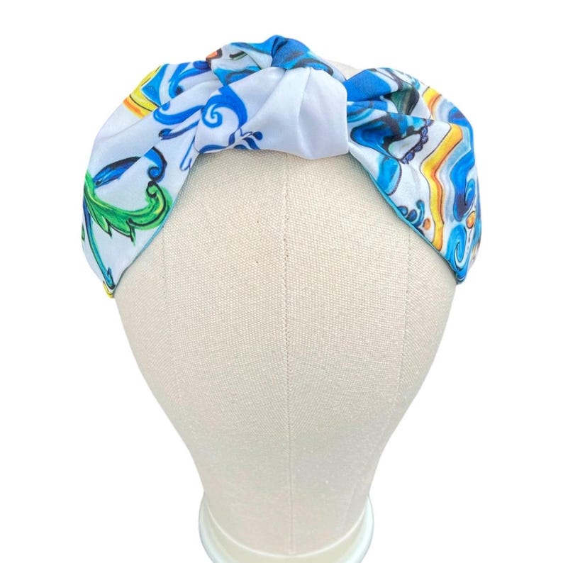 May include: A white headband with a blue, yellow, and green patterned design. The headband is tied in a knot on top of the head.
