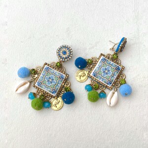 May include: A pair of blue and green statement earrings with a square tile design, gold accents, and blue and green pom-poms.