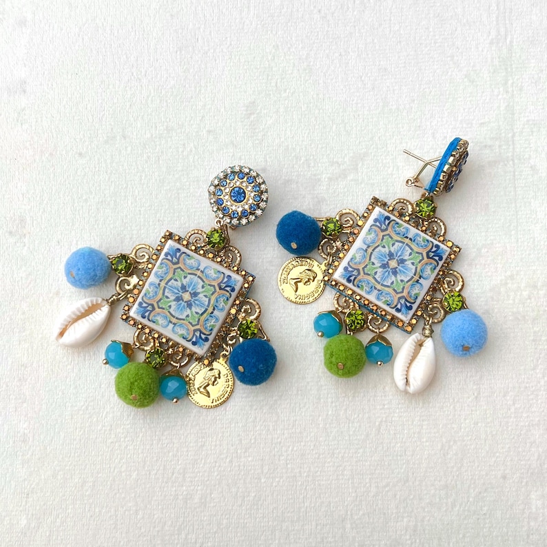 May include: A pair of blue and green statement earrings with a square tile design, gold accents, and blue and green pom-poms.