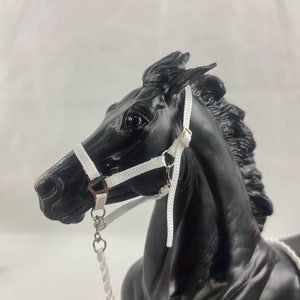 May include: A black horse figurine with a white halter and lead rope. The halter has silver-colored hardware and is secured around the horse's head. The horse's mane is flowing, and the figurine is set against a white background.