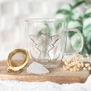 May include: A clear glass double-walled mug with a gold design of a moth and crescent moon. The mug is sitting on a wooden surface with a gold tea strainer and a white crystal.