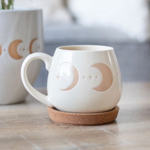 May include: White ceramic mug with a cork coaster. The mug features a design of two crescent moons and stars in a light peach colour.