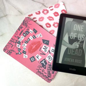 May include: A black Kindle e-reader showing the book "One of Us Is Dead" by Jeneva Rose, beside two decorative pink and white coasters. One coaster has a lip print design, and the other reads "Personalized by My Three".