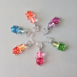 May include: A collection of colorful turtle-shaped earrings. Each earring features a small, translucent turtle charm in shades of blue, orange, pink, purple, and green. The turtle shells have a contrasting red outline. The earrings are connected to clear, plastic clip-on attachments.