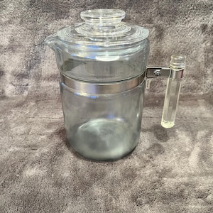May include: A clear glass coffee pot with a lid and a handle. The pot has a silver metal band around the middle and a clear plastic handle. The coffee pot is sitting on a gray surface.