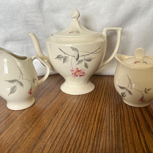May include: A cream-colored tea set featuring a teapot, creamer, and sugar bowl. Each piece is adorned with a delicate floral design of pink roses and gray leaves. The set is made of ceramic and sits on a wooden surface.