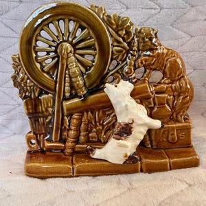 May include: A brown ceramic figurine depicting a spinning wheel, a bear, and a dog. The spinning wheel is in the center, with the bear on the right and the dog in front. The figurine has a glossy finish.